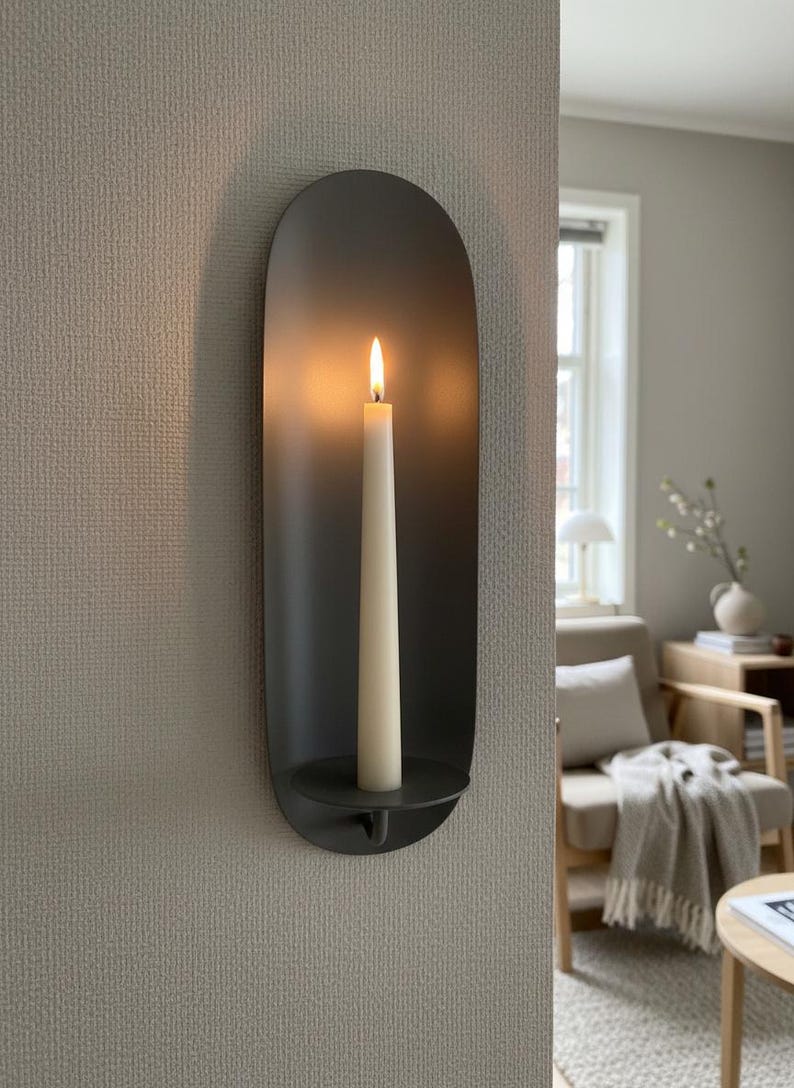 May include: A dark gray wall sconce with a lit, off-white candle. The sconce has a rounded, elongated shape and a small, circular base to hold the candle. The flame glows brightly against the dark background.