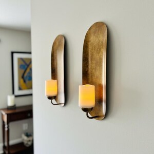 May include: Two wall sconces with a gold finish, each holding a battery-operated candle. The sconces have a curved, elongated design. The candles emit a warm, inviting glow, creating a cozy ambiance. The sconces are mounted on a light gray wall.