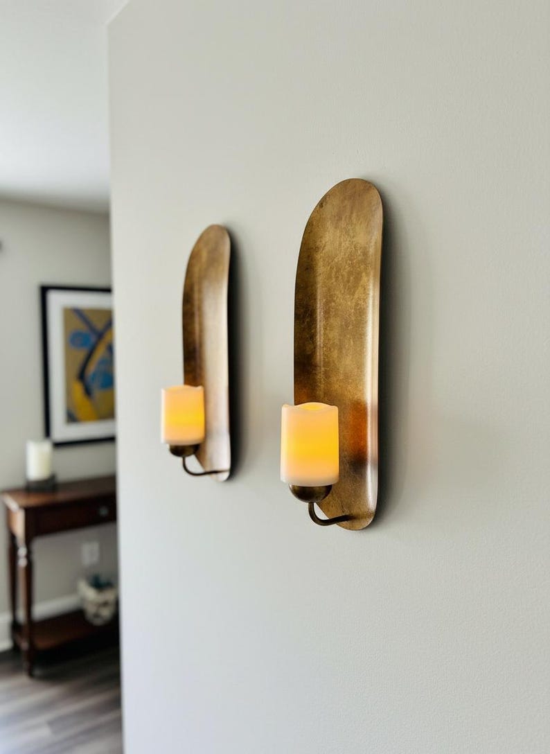 May include: Two wall sconces with a gold finish, each holding a battery-operated candle. The sconces have a curved, elongated design. The candles emit a warm, inviting glow, creating a cozy ambiance. The sconces are mounted on a light gray wall.