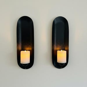 May include: Two black wall sconces with a matte finish, each holding a white pillar candle. The sconces have an oval shape with a slight gradient, and the candles emit a warm, inviting glow. Home decor.