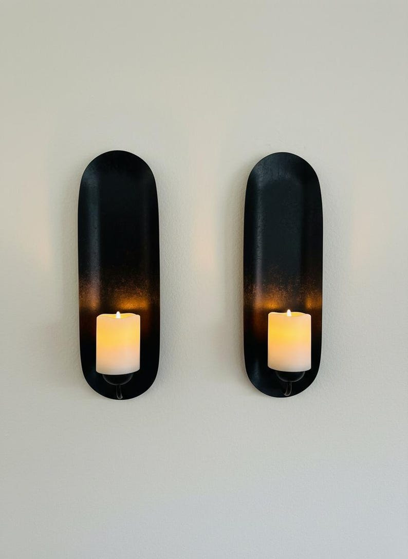 May include: Two black wall sconces with a matte finish, each holding a white pillar candle. The sconces have an oval shape with a slight gradient, and the candles emit a warm, inviting glow. Home decor.