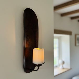 May include: A dark brown wall sconce with a faux candle. The sconce has a long, curved backplate and a small, curved arm that holds the candle. The candle is white with a flickering yellow light.