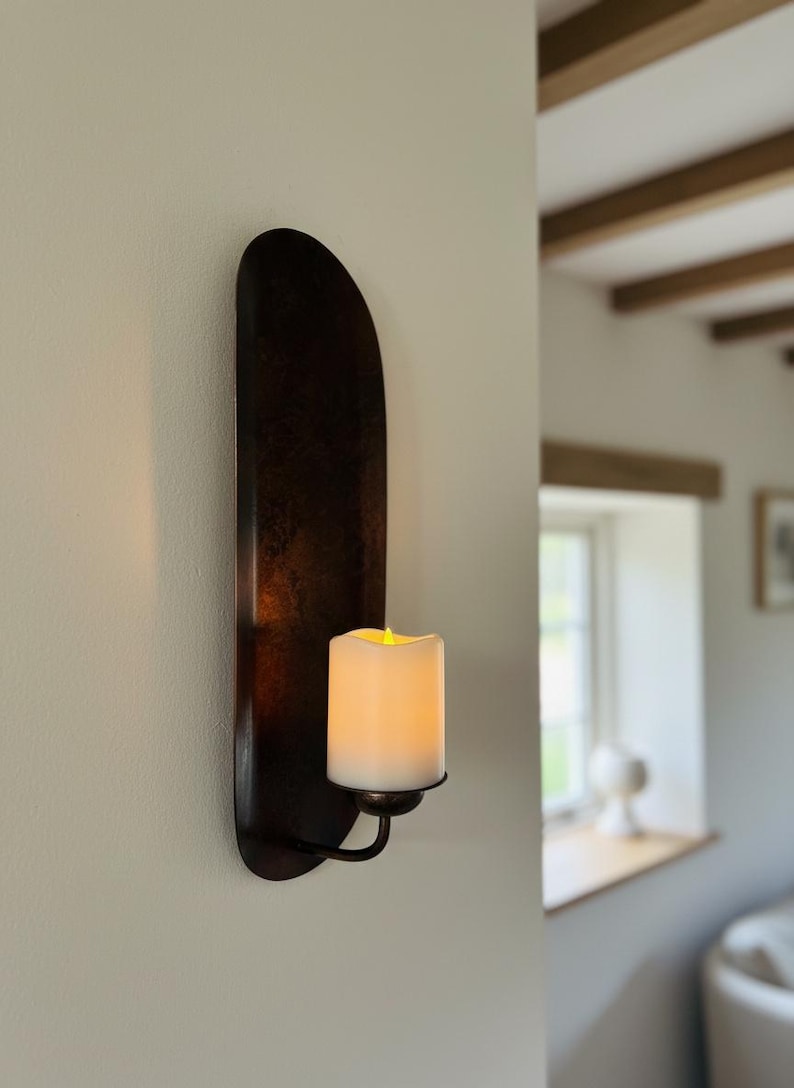 May include: A dark brown wall sconce with a faux candle. The sconce has a long, curved backplate and a small, curved arm that holds the candle. The candle is white with a flickering yellow light.