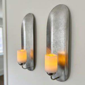 May include: Two wall-mounted candle holders with a brushed metal finish. Each holder features a curved, elongated backplate and a small, curved arm holding a cylindrical, battery-operated candle with a flickering yellow light.