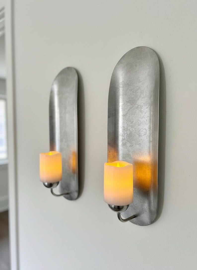 May include: Two wall-mounted candle holders with a brushed metal finish. Each holder features a curved, elongated backplate and a small, curved arm holding a cylindrical, battery-operated candle with a flickering yellow light.