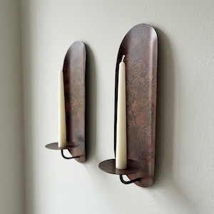 May include: Two wall-mounted candle holders with a rustic, copper-colored finish. Each holder features a tall, off-white candle. The holders have a curved, elongated design with a circular base to hold the candle.