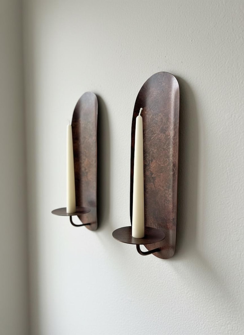 May include: Two wall-mounted candle holders with a rustic, copper-colored finish. Each holder features a tall, off-white candle. The holders have a curved, elongated design with a circular base to hold the candle.