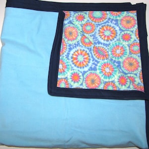 Puede incluir: Una manta azul claro con un borde azul marino. La manta presenta un estampado tie-dye en tonos naranja, rosa, verde y azul. La manta está doblada, mostrando la sección estampada.