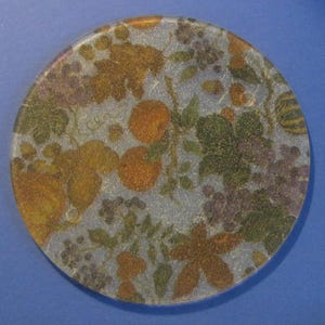 May include: A round, clear glass coaster with a fruit and foliage design. The coaster features oranges, grapes, and leaves in shades of gold, green, and purple. The design is set against a light blue background.