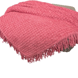 May include: A coral-colored throw blanket with a textured, woven pattern and fringed edges. The blanket is draped over a gray surface, creating a cozy and inviting aesthetic. Ideal for home decor and adding warmth.