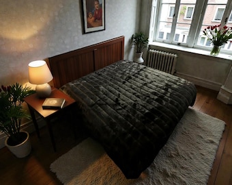 May include: A bedroom with a bed covered in a dark grey, textured quilt. A bedside table with a lamp and a book is visible. A white rug is on the floor. A window with a radiator and flowers in a vase is in the background.