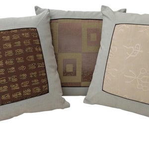 May include: Three decorative throw pillows. Each pillow has a light gray linen-like fabric with a square center panel. The center panels feature different patterns: brown hieroglyphs, geometric squares, and a cream-colored floral design.