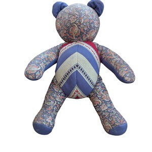 Puede incluir: Un oso de peluche hecho a mano con un estampado de cachemira en tonos azules, naranjas y rojos. El pecho del oso presenta un diseño de chevrón en azul, blanco y rojo. El oso tiene orejas y patas azules.