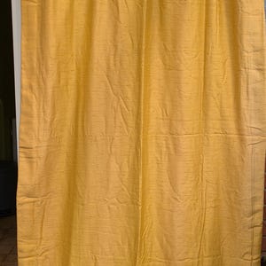 Pode incluir: Uma cortina amarelo-dourado feita de um tecido texturizado. A cortina está pendurada numa barra e é franzida na parte superior. O tecido tem um brilho subtil e um aspeto ligeiramente enrugado.