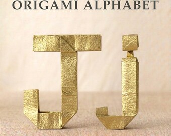 Könnte beinhalten: Der Text ORIGAMI ALPHABET steht über einem großen J und einem kleinen j, die beide aus goldfarbenem Papier mit einer strukturierten, gefalteten Oberfläche gefertigt sind. Die Buchstaben sind mit Origami-Techniken und sichtbaren Falten konstruiert.