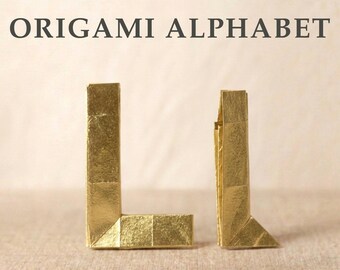 May include: The text ORIGAMI ALPHABET is displayed above two gold-colored paper folded letters. The letter L is on the left, and a smaller, narrower letter L is positioned to its right. Both letters feature sharp, geometric folds and a metallic finish.