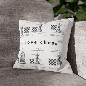 May include: White square pillow with a chess theme. The pillow features chess pieces, diagrams, and the text "i love chess". The design includes the names of the chess pieces and their movements. The pillow is on a gray sofa.