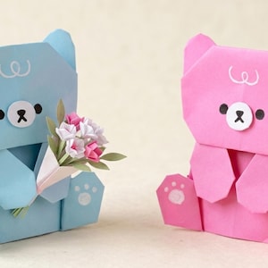 Könnte beinhalten: Zwei Origami-Bärenfiguren, einer hellblau und einer rosa, werden gezeigt. Der blaue Bär hält einen Papierstrauß aus rosa und weißen Blumen. Beide Bären haben schwarze Augen, eine weiße Nase und Pfotenabdrücke auf ihren Füßen.