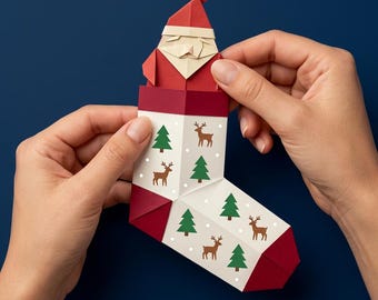Könnte beinhalten: Eine Origami-Weihnachtsmannfigur, die aus einem weißen Papierstrumpf hervorschaut. Der Strumpf ist mit einem wiederkehrenden Muster aus grünen Tannenbäumen und braunen Rentiersilhouetten bedruckt. Die Spitze und das Ende sind dunkelrot.