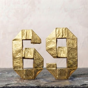 May include: Two three-dimensional numbers, six and nine, crafted from gold-coloured material with a textured, folded surface. The numbers are upright and display a geometric, angular construction with sharp edges and a metallic, reflective finish.