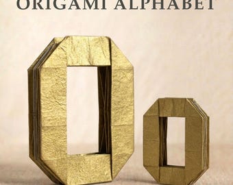 Könnte beinhalten: Zwei goldfarbene Origami-Buchstaben in Form eines O, einer groß und einer klein, nebeneinander dargestellt. Der Text ORIGAMI ALPHABET ist oben in Dunkelgrau gedruckt. Die Buchstaben haben eine gefaltete Papierstruktur mit scharfen Kanten.