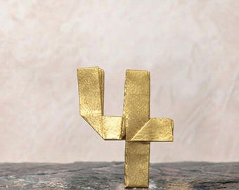 Könnte beinhalten: Eine goldfarbene Ziffer Vier aus gefaltetem Material mit metallischem Glanz und scharfen, kantigen Falten. Die Ziffer steht aufrecht und zeigt eine strukturierte Oberfläche mit sichtbaren Knicken, die eine geometrische Form der Zahl Vier bilden.