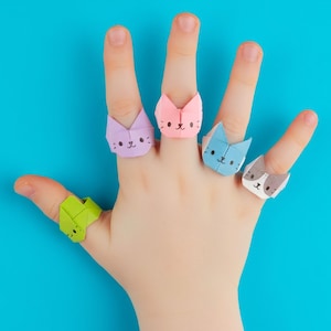 Op de afbeelding: Vijf origami papieren ringen in de vorm van kattenkoppen in diverse kleuren, waaronder limoengroen, lichtpaars, zachtroze, lichtblauw en wit met grijze vlekken. Elke ring is voorzien van een handgetekend kattengezicht met ogen en snorharen.