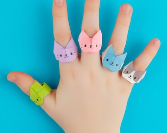 Könnte beinhalten: Fünf Origami-Papierringe in Form von Katzenköpfen in verschiedenen Farben, darunter Limettengrün, Helllila, zartes Rosa, Hellblau und Weiß mit grauen Flecken. Jeder Ring zeigt ein handgezeichnetes Katzengesicht mit Augen, Nase und Schnurrhaaren.