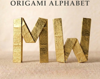 May include: Two gold-colored letters, M and W, crafted from folded paper with a metallic, crinkled texture. The letters stand upright, and the text ORIGAMI ALPHABET is printed in dark gray capital letters above them on a light background.