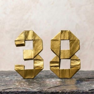 Op de afbeelding: Twee driedimensionale cijfers, drie en acht, gemaakt van metallic goudkleurig papier met zichtbare vouwlijnen en scherpe geometrische randen. De cijfers staan rechtop en vertonen een reflecterend oppervlak en een gestructureerd, hoekig origami-ontwerp.