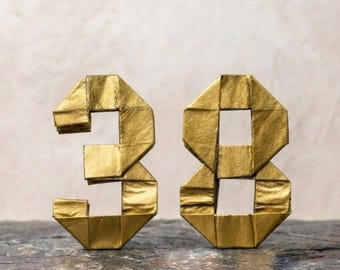 Könnte beinhalten: Zwei dreidimensionale Zahlen, drei und acht, gefertigt aus metallisch goldenem Papier mit sichtbaren Faltkanten und scharfen geometrischen Rändern. Die Zahlen stehen aufrecht und zeigen eine reflektierende Oberfläche sowie ein strukturiertes Origami-Design.