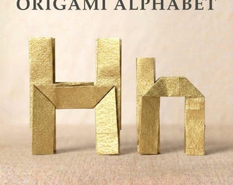 Könnte beinhalten: Der Großbuchstabe H und der Kleinbuchstabe h sind aus goldfarbenem Papier in Origami-Technik gefaltet. Der Text ORIGAMI ALPHABET ist in Dunkelgrau über den beiden goldenen Papierbuchstaben gedruckt, die sichtbare Faltkanten und eine Textur aufweisen.
