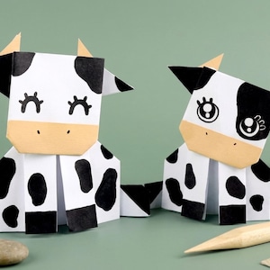 Può includere: Due figure di mucca origami, una con gli occhi chiusi e l'altra con gli occhi aperti, sono affiancate. Le mucche sono bianche con macchie nere, orecchie nere e facce color beige. Lo sfondo è verde.