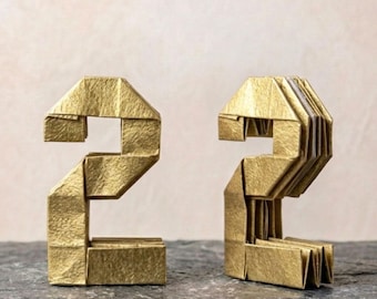 Könnte beinhalten: Zwei goldfarbene Origami-Zahlen in Form der Ziffer Zwei, gefertigt aus strukturiertem Papier. Die linke Zahl ist von vorne zu sehen, während die rechte Zahl schräg steht und die geschichtete, gefaltete Papierkonstruktion mit Akkordeon-Optik zeigt.
