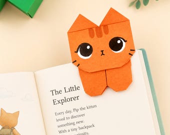 Könnte beinhalten: Ein orangefarbenes Origami-Lesezeichen aus Papier in Form eines Kätzchens mit großen schwarzen Augen, einer kleinen Nase und drei Streifen auf der Stirn. Das Kätzchen ist so geformt, dass es auf die Ecke einer Buchseite passt, die den Text The Little Explorer zeigt.