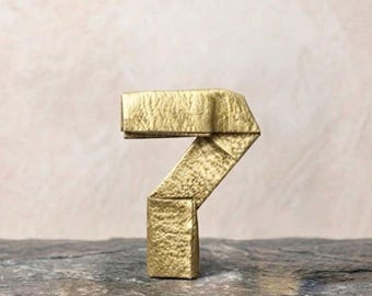 Könnte beinhalten: Eine dreidimensionale Ziffer Sieben aus goldfarbenem Material mit einer texturierten, zerknitterten Oberfläche. Die Ziffer weist scharfe, gefaltete geometrische Kanten auf und steht aufrecht, wobei sie ein metallisches Finish zeigt.