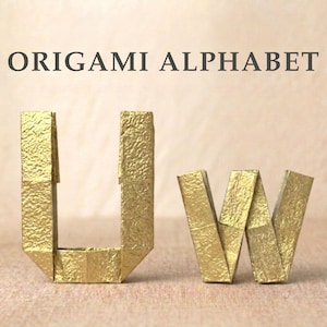 Puede incluir: Dos letras de origami de color dorado, U y W, con una superficie metálica texturizada. Las letras se muestran en posición vertical con el texto ORIGAMI ALPHABET impreso en letras mayúsculas de color gris oscuro sobre un fondo claro y neutro.