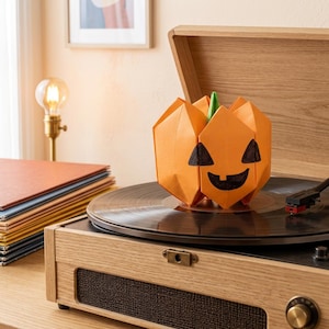 May include: An orange origami pumpkin with a green stem and a hand-drawn black jack-o'-lantern face sits on the black platter of a light wood record player. The paper pumpkin features sharp geometric folds and a wide, smiling mouth.