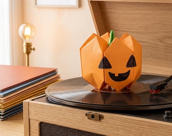 Könnte beinhalten: Ein orangefarbener Origami-Kürbis mit grünem Stiel und einem handgezeichneten schwarzen Jack-o'-Lantern-Gesicht steht auf dem schwarzen Plattenteller eines hellen Holz-Plattenspielers. Der Papierkürbis hat scharfe geometrische Falten.