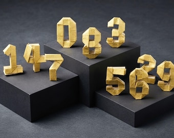 Könnte beinhalten: Eine Sammlung von zehn dreidimensionalen Zahlen aus goldfarbenem Papier, die die Ziffern null bis neun darstellen. Jede Zahl ist mit sichtbaren Faltkanten und geometrischen Facetten konstruiert und steht aufrecht wie eine Papierskulptur.