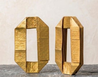 Könnte beinhalten: Zwei rechteckige Creolen aus metallisch goldenem Papier mit gefalteter Origami-Struktur. Jeder Ohrring verfügt über eine hohle Mitte und eine geometrische, mehrschichtige Konstruktion mit scharfen, kantigen Formen und einer deutlich geknitterten Oberfläche.