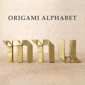 Może przedstawiać: Tekst ORIGAMI ALPHABET jest wydrukowany w kolorze ciemnoszarym nad trzema złotymi papierowymi literami złożonymi w kształty małych liter m, n i u. Każda litera posiada ostre, geometryczne zagięcia charakterystyczne dla sztuki składania papieru.
