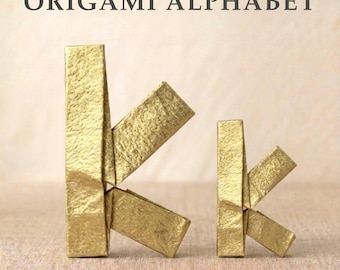 Könnte beinhalten: Der Text ORIGAMI ALPHABET steht über einem großen Buchstaben K und einem kleinen Buchstaben k, die beide aus goldfarbenem Papier mit einer strukturierten, gefalteten Oberfläche gefertigt sind. Die Buchstaben stehen aufrecht und zeigen die Falttechnik.