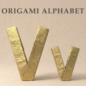 May include: A large and a small letter V crafted from gold-colored paper with a textured, crinkled finish. The text ORIGAMI ALPHABET is printed in dark gray capital letters above the two gold paper letters, which stand upright on a flat surface.