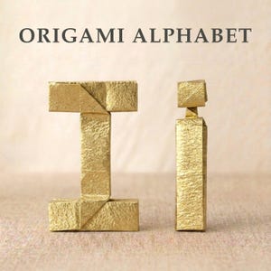 May include: The text ORIGAMI ALPHABET is displayed above two gold-colored paper folded letters. The uppercase letter I is on the left, and the lowercase letter i is on the right. Both letters feature a textured, metallic finish and sharp folds.