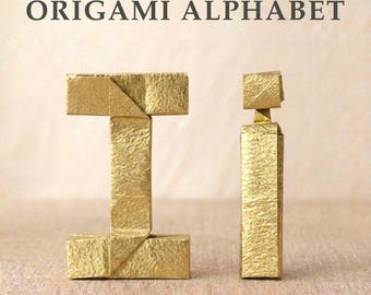 Könnte beinhalten: Der Text ORIGAMI ALPHABET steht über zwei aus goldfarbenem Papier gefalteten Buchstaben. Der Großbuchstabe I befindet sich links und der Kleinbuchstabe i rechts. Beide Buchstaben haben eine strukturierte, metallische Oberfläche und scharfe Falten.
