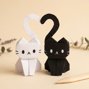 May include: Two origami cat figures, one white and one black, sit side-by-side. Their tails curve upwards, forming a heart shape. Each cat has a simple face with black whiskers and eyes. The background is a soft beige.