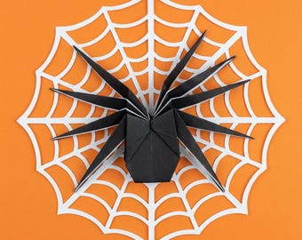 Könnte beinhalten: Eine schwarze Origami-Spinne mit acht spitzen Beinen, die in der Mitte eines weißen, ausgestanzten Spinnennetzes aus Papier platziert ist. Die Spinne und das Netz sind vor einem einfarbigen orangefarbenen Hintergrund angeordnet.