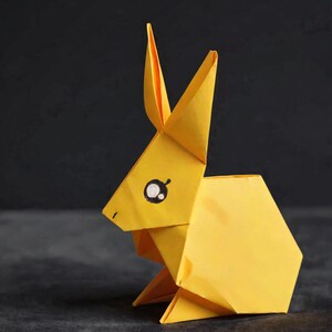 Puede incluir: Una figura de conejo de origami de color amarillo brillante hecha de papel plegado. El conejo presenta dos orejas largas y puntiagudas, una estructura corporal geométrica y un ojo visible con un contorno negro y un pequeño detalle blanco.