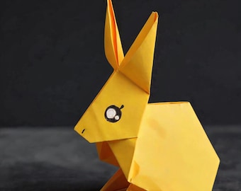 Könnte beinhalten: Eine hellgelbe Origami-Hasenfigur aus gefaltetem Papier. Der Hase hat zwei lange, spitze Ohren, einen geometrischen Körperbau und ein einzelnes sichtbares Auge, das mit einer schwarzen Umrandung und einem kleinen weißen Lichtpunkt dargestellt ist.